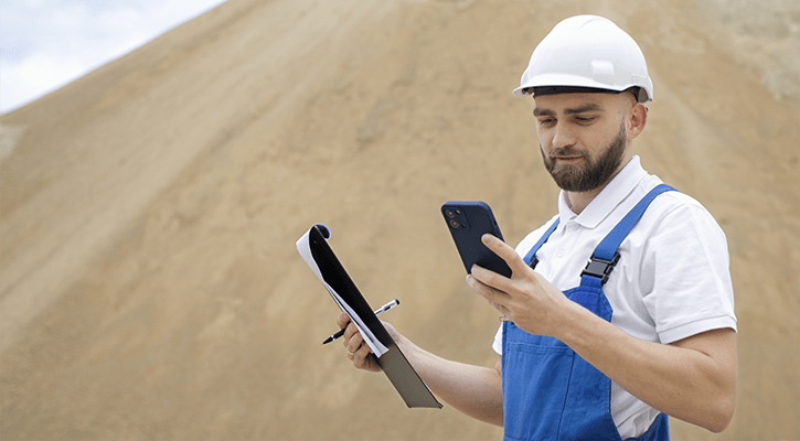 A site worker in a hard hat checking a phone on open ground